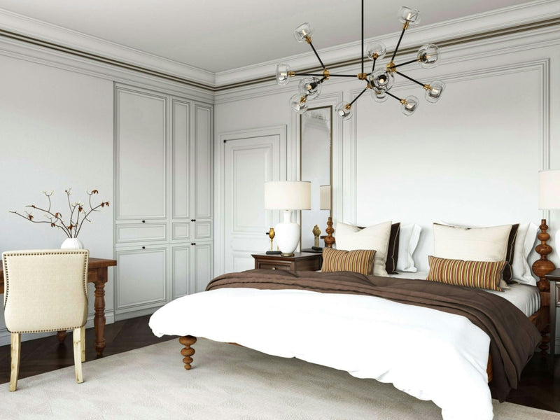 A modern bedroom showcases the Ophelia Bed - Tuscany by James+James, adorned with chic white and brown bedding. Flanking the bed are two wooden nightstands, each topped with a matching lamp. Overhead hangs a contemporary chandelier, providing stylish illumination to the room, which features elegant white paneled walls and built-in wardrobes. The space is further enhanced by a wooden desk paired with a cream chair and accessorized with a vase containing minimalist bare branches.