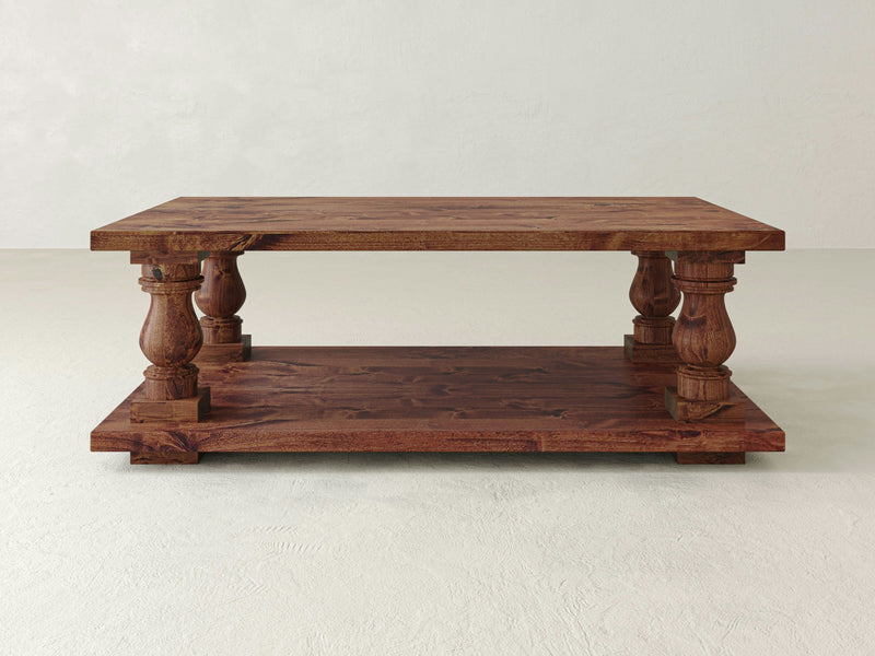 The James+James Vivien Coffee Table showcases a refined design with intricate, ornate legs and a rectangular shape. This wooden table exemplifies artisanal craftsmanship, featuring a lower shelf and a natural wood finish that highlights its rich grain and sturdy build. It is set against a plain, light-colored wall and floor background.