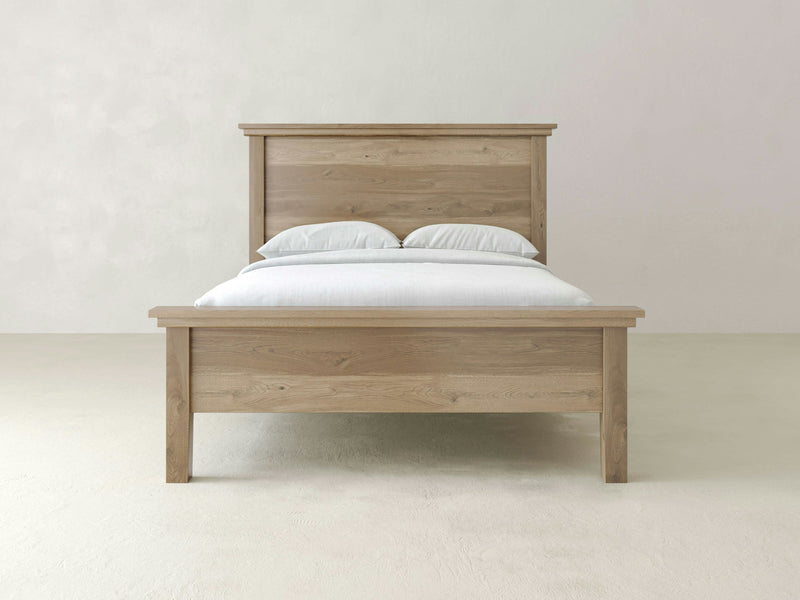 The Ivy Bed - Barn Wood by James+James is a wooden bed frame with a high headboard and low footboard, both featuring a simple, clean design. The bed is crafted from light, natural-colored wood and is neatly made with white linens and two pillows against a plain, light-colored background.