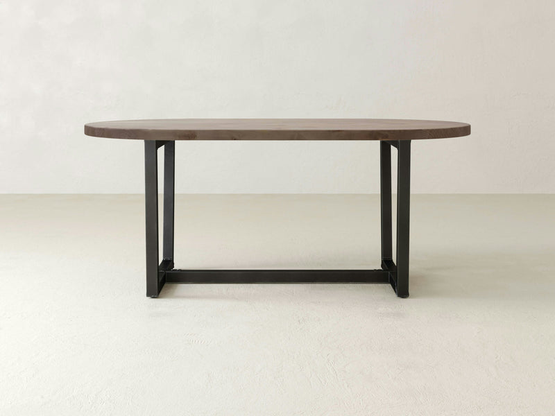 The Trapezoid Oval Dining Table - Barn Wood by James+James stands in an empty, light-colored room. Its minimalist design featuring an oval barn wood top and black metal legs is highlighted against the plain backdrop, emphasizing its simple and modern aesthetic.