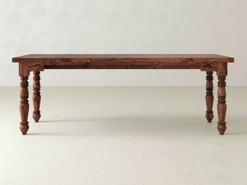 The Turned Leg Desk - Tuscany by James+James is a rectangular wooden table with a simple, rustic design, featuring four intricately carved legs. The desk is set against a plain, light-colored background.