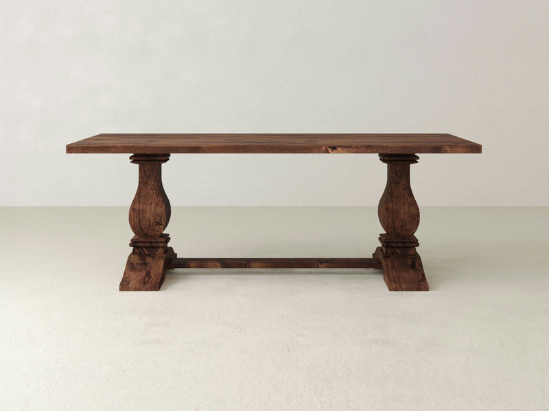 The Heirloom Dining Table - Tuscany by James+James features a rustic wooden design with a flat rectangular top and two ornate pedestal legs connected by a horizontal support. This table showcases a natural wood grain finish and is perfectly set on a light, neutral tiled floor against a plain, light-colored wall.