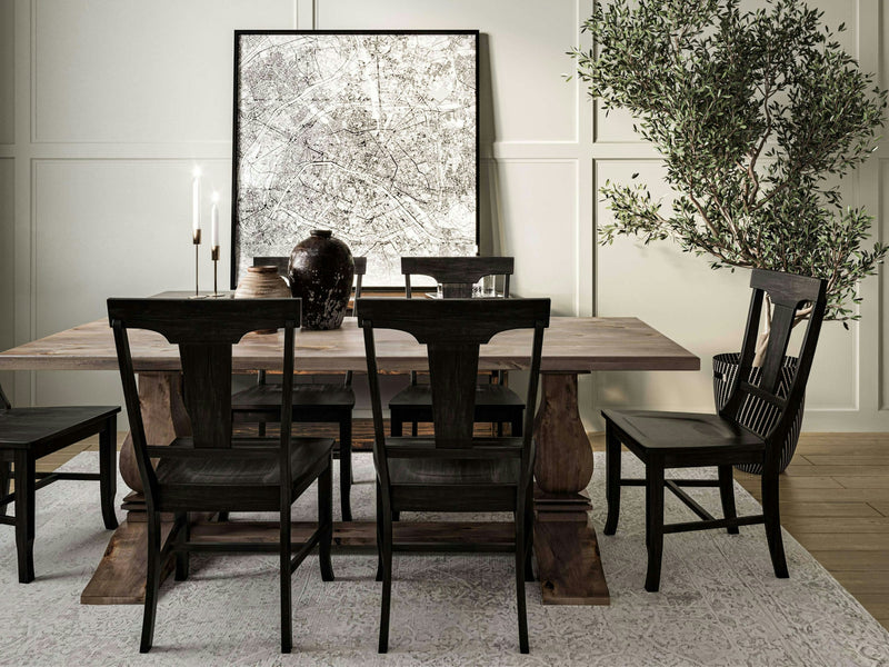 A stylish dining room featuring the Heirloom Dining Table in barn wood by James+James, accompanied by six black chairs. The table is adorned with large ceramic vases and two lit candles. A textured artwork leans against the wall, and a leafy green tree adds a natural touch to the space. The floor has a light-colored rug.