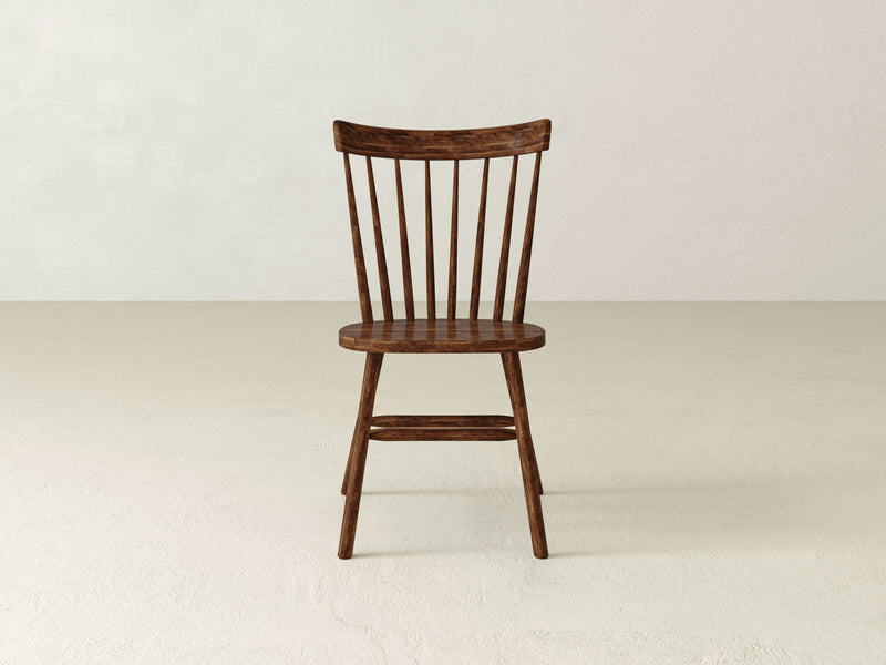 A Rustic Windsor Dining Chair - Tuscany by James+James, with a darker wood finish, is centered in the image. The chair features a classic spindle back design and four straight legs. The background is a plain, off-white wall with a light-colored floor.