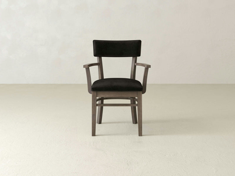 The Everly Arm Dining Chair by James+James stands against a plain, light-colored wall and floor. This comfortable armchair, featuring a single wooden frame with a dark cushioned seat and backrest, presents clean lines and armrests in a minimalist design that adds a touch of mid-century modern elegance.