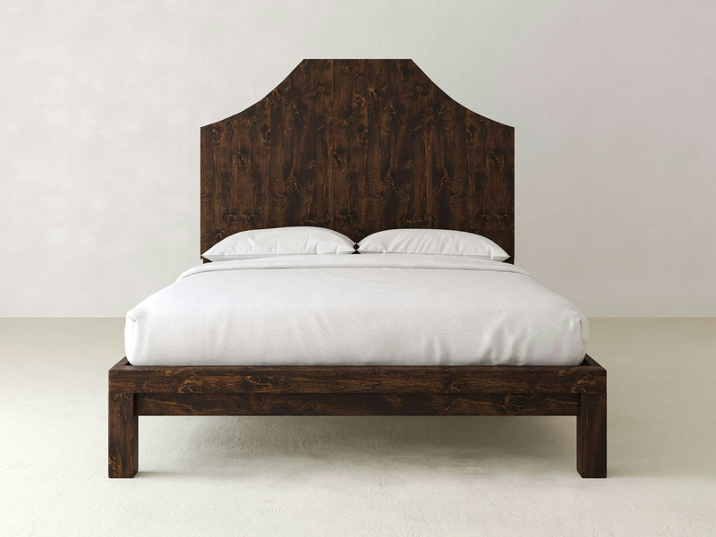 A Megan Bed in Tobacco finish from James+James, featuring a tall and ornate headboard, stands against a plain, light-colored wall. The bed is neatly made with white bedding and two white pillows, creating a clean and minimalistic look.