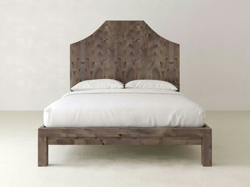 The Megan Bed - Barn Wood by James+James, featuring a minimalist design with a tall, arched headboard, is centered against a plain, light-colored wall. The bed is dressed in white bedding with two pillows and a neatly tucked blanket. The frame and headboard boast a rustic, unpolished finish.