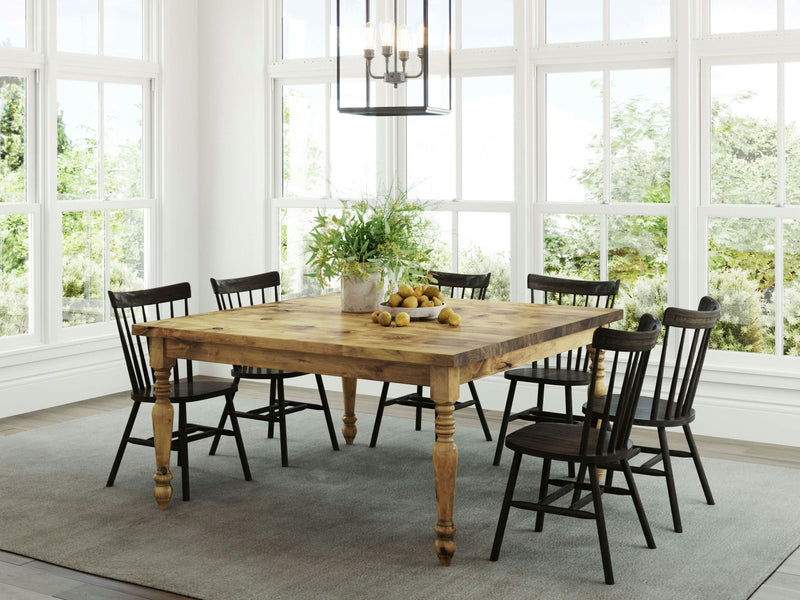 A bright dining area features a James+James French Country Square Dining Table in Harvest Wheat, surrounded by six dark wooden chairs. A vase with green foliage and a bowl of lemons sit on the table, while large windows in the background allow natural light to flood the room, showcasing the outdoor greenery.