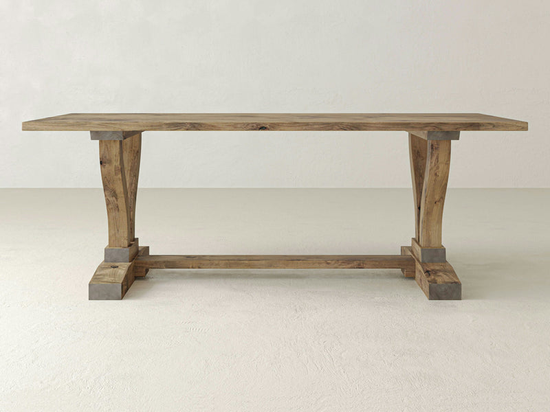 The Vera Dining Table - Harvest Wheat by James+James is a rectangular wooden dining table with a natural finish. It features a sturdy trestle base with two thick, tapered legs connected by a horizontal support beam. The setting is a minimalistic room with light-colored walls and a light floor.