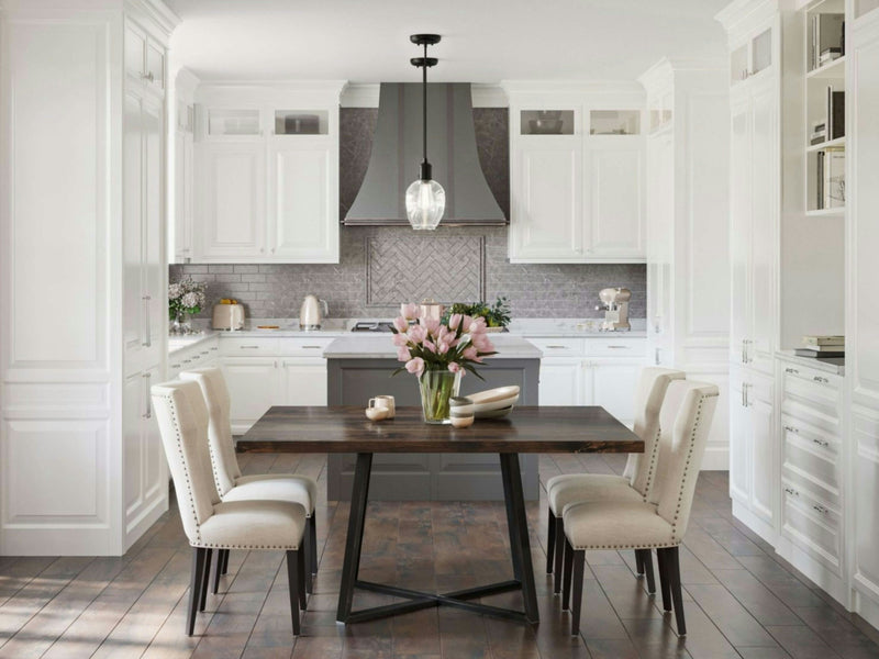 A modern kitchen with white cabinetry, gray brick backsplash, and stainless steel appliances. In the center is the Watson Square Dining Table from James+James in a rich tobacco finish, surrounded by four beige upholstered chairs and topped with a vase of pink tulips. A black pendant light hangs above the table.