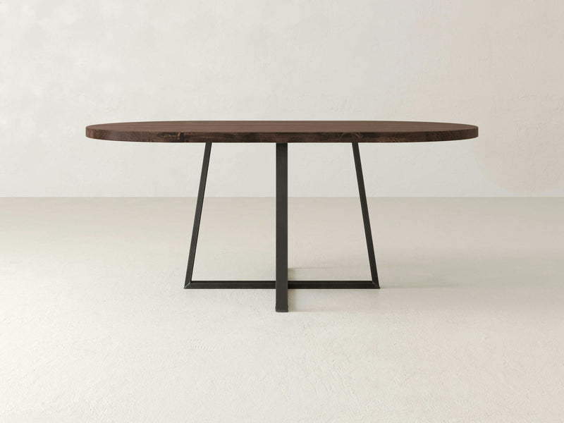 The Watson Oval Dining Table by James+James features a minimalist design with a dark tobacco wooden top and a sleek black metal base. Its simple, clean lines and contemporary style stand out beautifully against a plain, light-colored background.