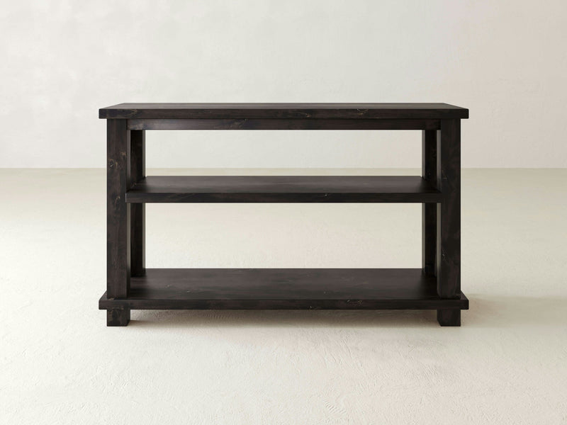 The Carter X Sofa Table in Charred Ember by James+James is a dark wooden console table with three tiers, standing on a light-colored floor against a light background. It features a simple, rectangular design with a sturdy base and open shelving for storage or display.