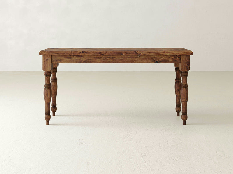 The Abigail Sofa Table - Tuscany by James+James features a rectangular top and four ornately carved legs. It is showcased on a light-colored floor against a plain, light-colored wall. The table's natural, rustic finish beautifully highlights its grain patterns.