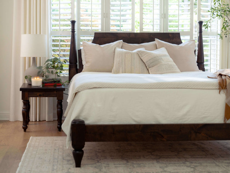 A cozy bedroom featuring a dark wooden bed with white and beige bedding, accompanied by a James+James Abigail End Table in Tobacco with a lamp, a plant, and a candle. Large windows with white blinds let natural light into the room, and light-colored curtains frame the windows.