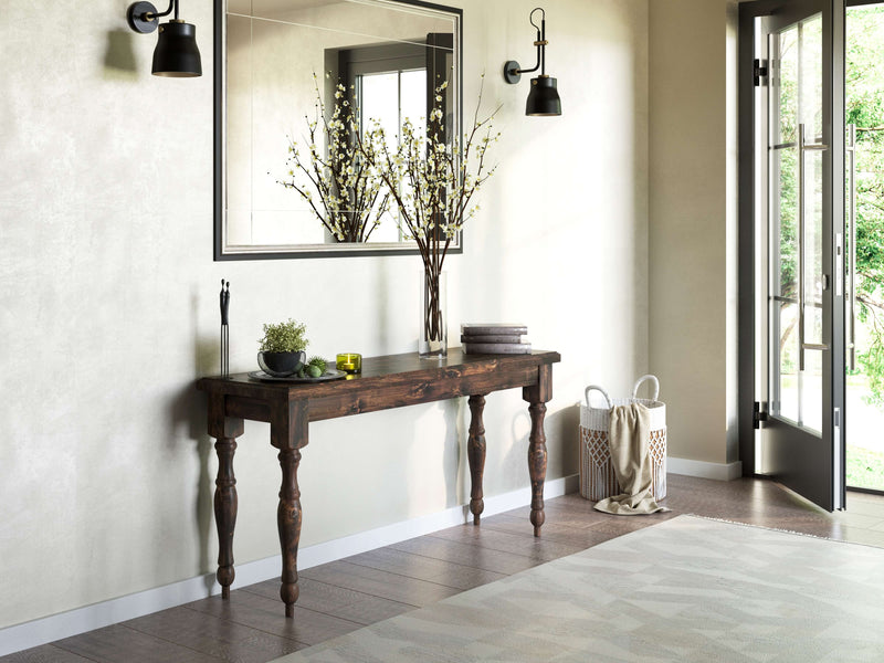 A minimalist entryway features the Abigail Sofa Table in Tobacco by James+James, topped with a vase of branches, a small black sculpture, a candle, and stacked books. Two black sconces flank a large mirror on the wall, and a woven basket sits near an open door leading outside.