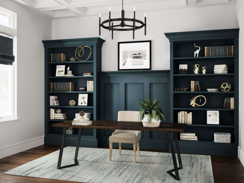 Elegant home office with a James+James Arkwright Desk in a rich tobacco finish and a cream chair centered on a patterned rug. Built-in teal bookshelves are arranged behind the desk, filled with books, decorative items, and a potted plant. A black modern chandelier hangs from the coffered ceiling.