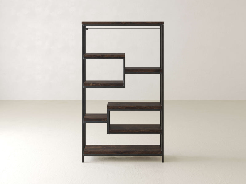 The James+James Arlo Shelf is a modern bookshelf with a geometric, zigzag design. It features multiple solid hardwood shelves framed in industrial steel, creating open spaces of varying heights and widths that are perfect for books or decor. The background consists of plain, light-colored walls and floors.