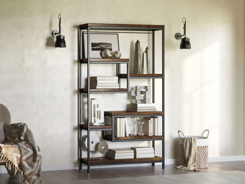 The Arlo Shelf by James+James showcases solid hardwood shelves with customizable stain finishes, adorned with books and decor like abstract sculptures and hourglasses. It's flanked by two wall-mounted black lamps, while a patterned blanket is draped over a chair against the soft beige wall.