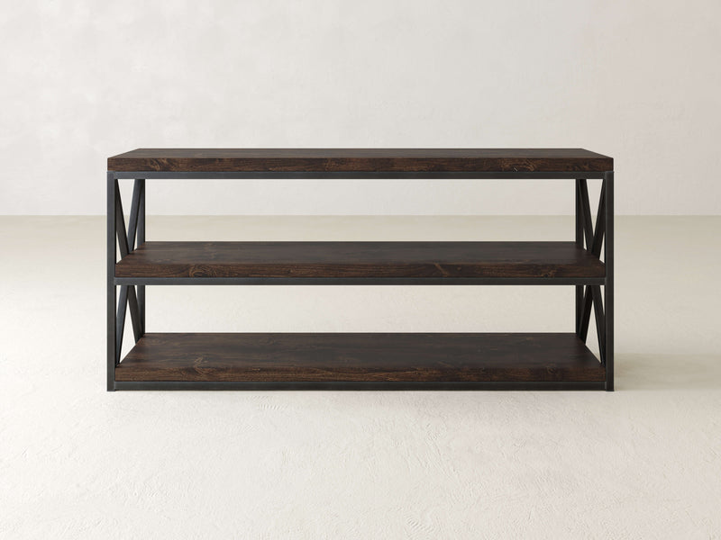 A minimalist dark wood console table from James+James, named the Boyd Shelf, offers three spacious shelves and an industrial steel frame with open sides featuring an X-pattern for added support. The stylish piece is set against a plain, neutral wall and floor.