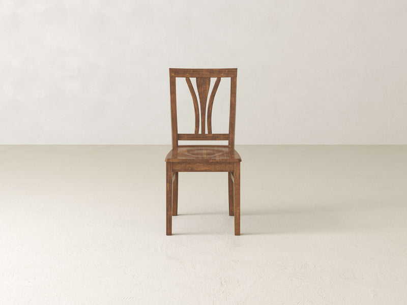 The Brooklyn Dining Chair - Tuscany by James+James, featuring an elegant design with intricately carved details on the backrest, stands alone on a plain, light-colored floor against a beige wall. The minimalistic scene highlights the chair's craftsmanship.