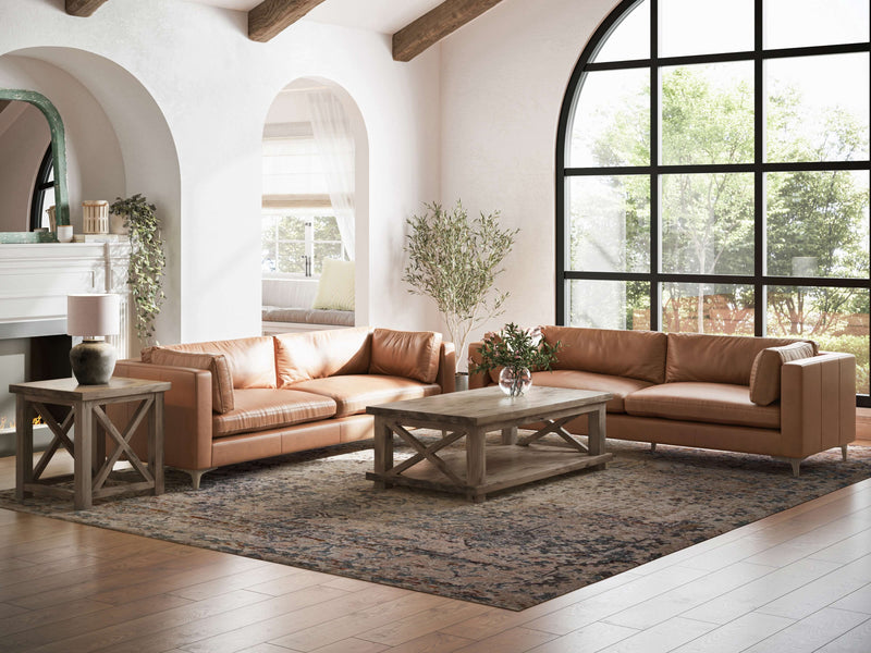 Bright living room with high, arched ceilings and large, black-framed windows. Two tan leather sofas are arranged in an L-shape around a rustic wooden coffee table. Nearby, a James+James Carter X Side Table - Barn Wood holds a lamp. An area rug covers the wooden floor.