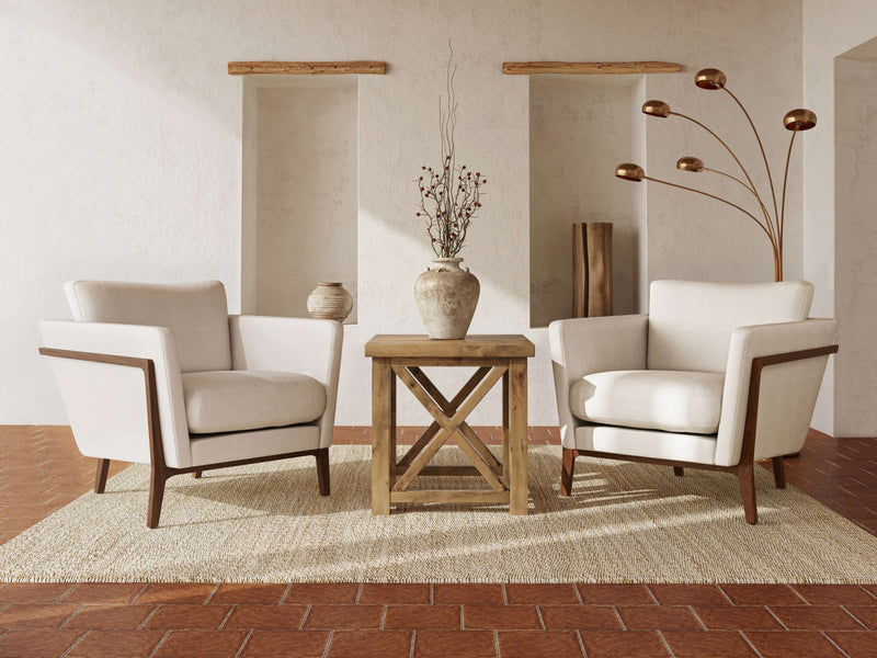 A modern living room with two beige armchairs flanking the Carter X Side Table in Harvest Wheat by James+James on a textured rug. Decorative vases, a minimalist floor lamp with spherical lights, and recessed wall niches complete the serene and elegant setting.