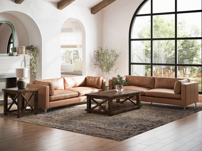 A spacious living room with large arched windows and white walls features two tan leather sofas, a rustic wooden coffee table, and a matching Carter X Side Table - Tobacco by James+James with a lamp. The room is decorated with potted plants and an ornate area rug on the wooden floor.