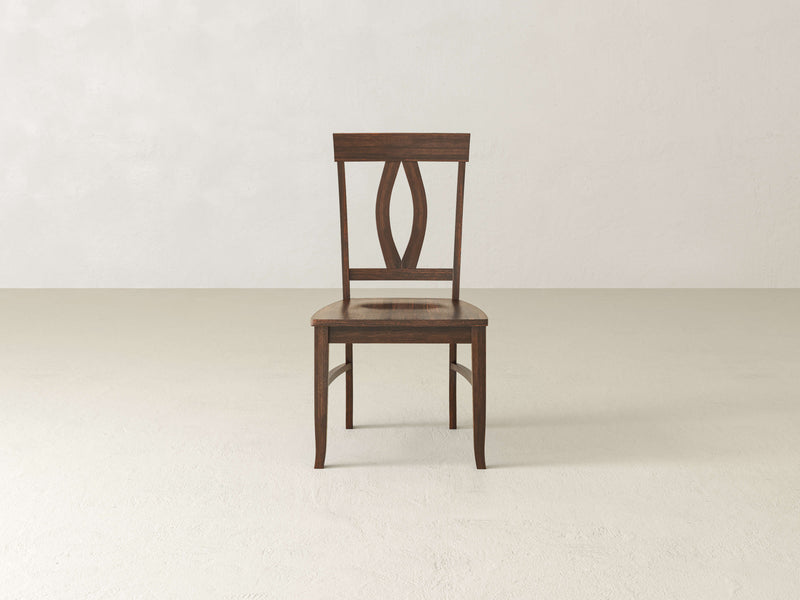 The Elizabeth Dining Chair - Tobacco by James+James features a simple, elegant design. This wooden chair boasts a dark brown finish, a slightly curved backrest with an openwork oval pattern in the center, and four straight legs. It is positioned on a beige floor against a light-colored minimalist wall.