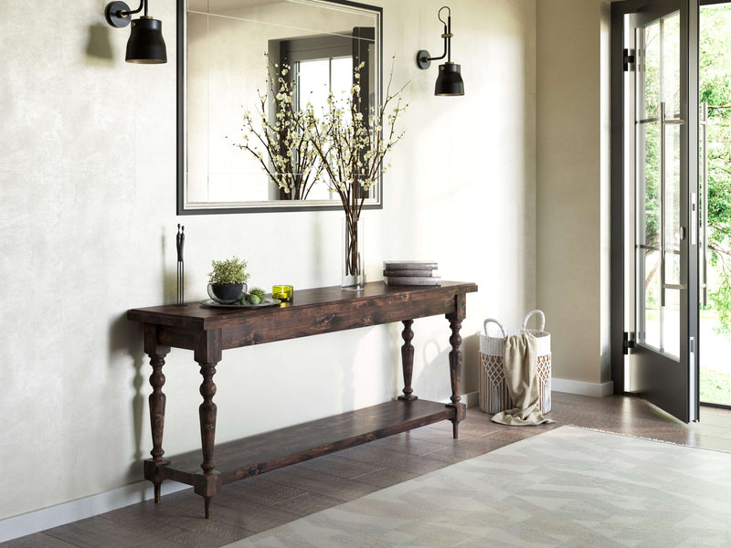 A rustic entryway features the Ella Sofa Table in Tobacco by James+James, adorned with a vase of branches, a potted plant, and stacked books. A large mirror with a simple wooden frame hangs above. Two black sconces flank the mirror. An open door reveals greenery outside.