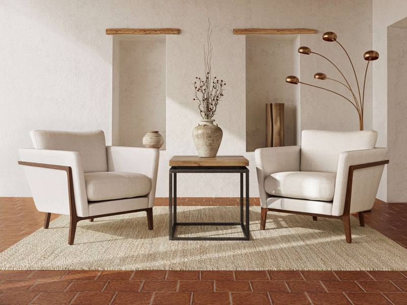 A cozy and minimalist sitting area features two beige armchairs facing each other, separated by the James+James Floating Top End Table in Harvest Wheat color placed on a textured beige rug. Behind the armchairs is a white textured wall with built-in niches displaying decorative vases and a tall floor lamp.