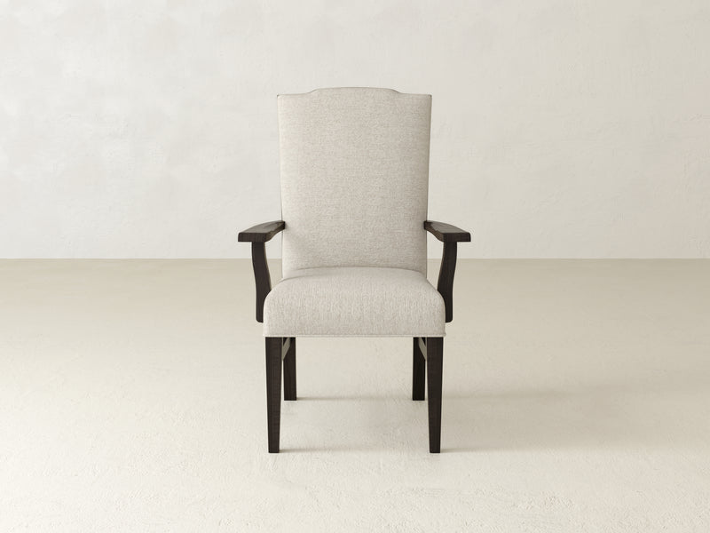 A Hadley Arm Dining Chair in Charred Ember by James+James stands against a light textured wall and floor. This chair features dark wooden legs and armrests, with a high back and a simple, elegant design.