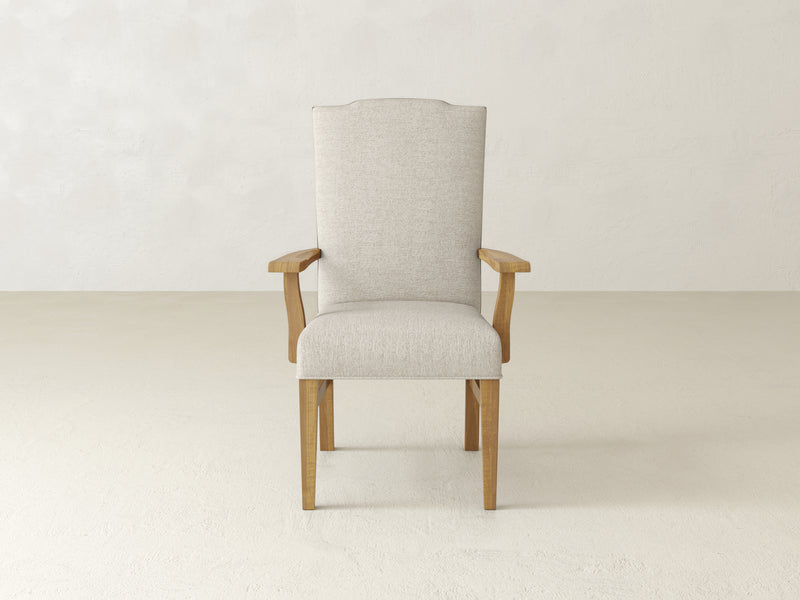 The Hadley Arm Dining Chair - Harvest Wheat by James+James is displayed in a minimalistic setting with a light, textured wall and floor, featuring beige upholstery and wooden arms and legs.