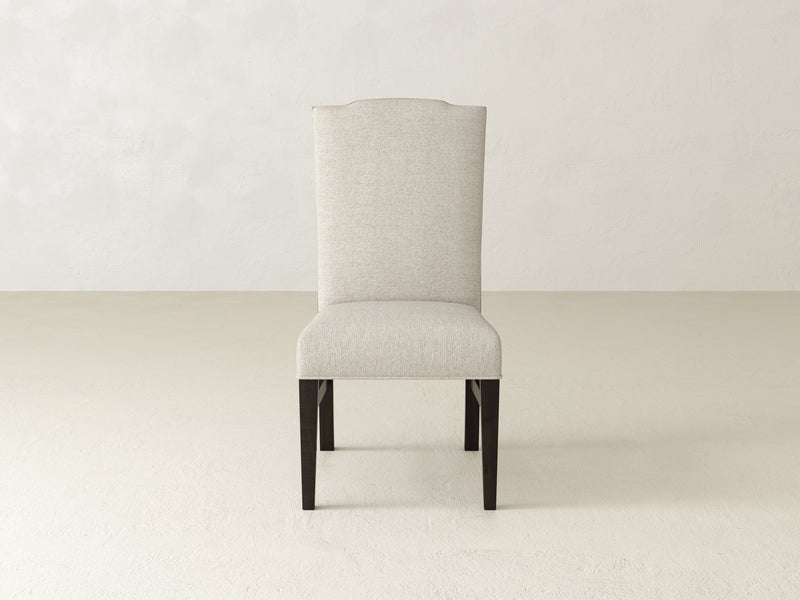 A beige Hadley Side Dining Chair by James+James, featuring a high backrest and dark wooden legs in Charred Ember, set on a light, neutral-colored floor against a plain wall.