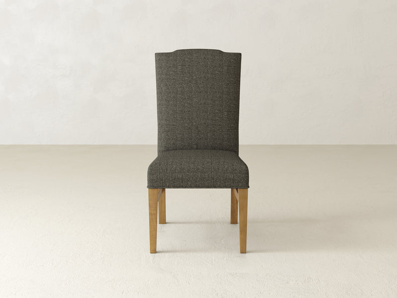 A Harvest Wheat Hadley Side Dining Chair by James+James, featuring a minimalist design with wooden legs and an upholstered high backrest, stands on a plain light-colored floor against a textured off-white wall.