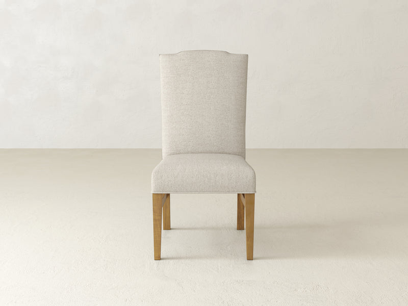 The Hadley Side Dining Chair - Harvest Wheat by James+James, featuring a beige upholstery, high backrest, and wooden legs, stands on a light-textured floor with a plain, light-colored wall in the background.