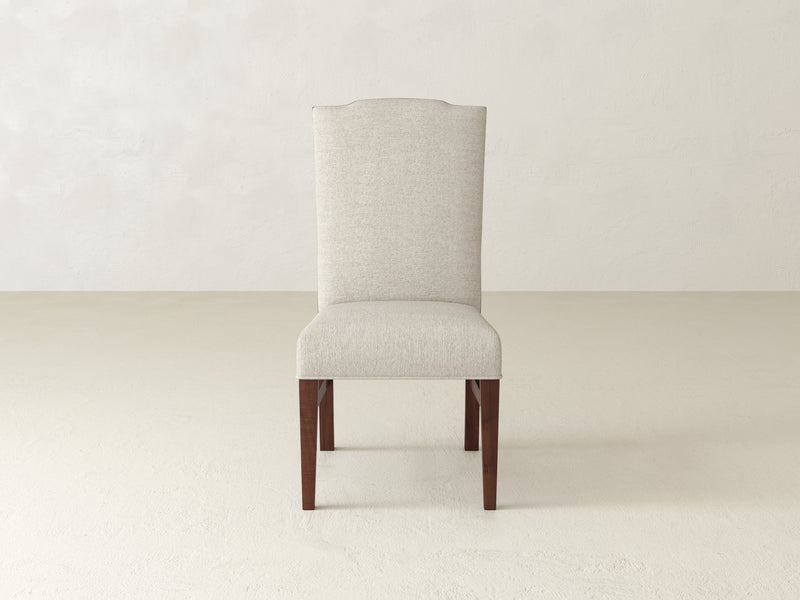 The Hadley Side Dining Chair - Tuscany from James+James, featuring a beige upholstery with a high backrest and wooden legs, is placed on a light-colored floor against a plain, light-colored wall.