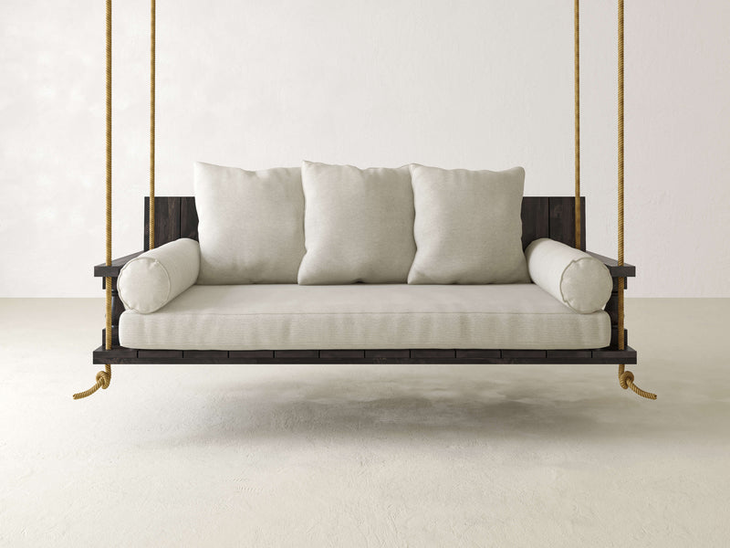 A James+James Maisie Porch Swing in Charred Ember, adorned with beige cushions, is suspended by four ropes in a minimalist, white-walled room with a concrete floor.