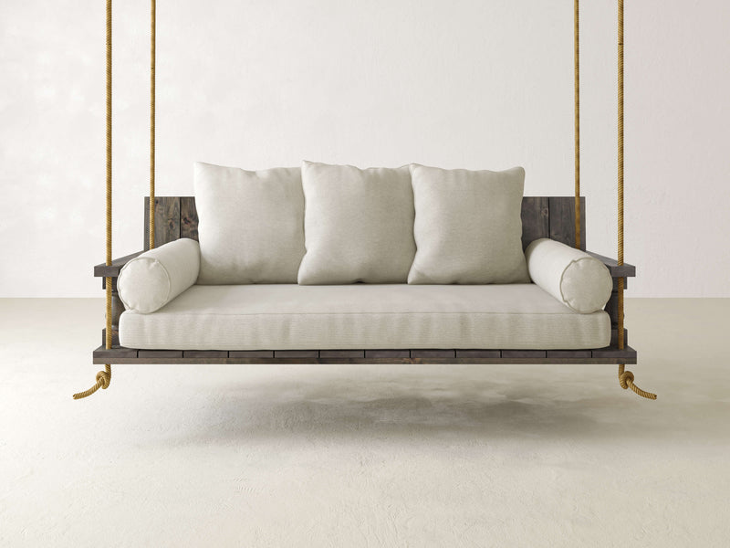 A minimalist James+James Maisie Porch Swing in deep grey, complete with three matching pillows, is suspended by thick ropes in a bright, neutral-toned room with white walls and floors, creating a serene and airy atmosphere.