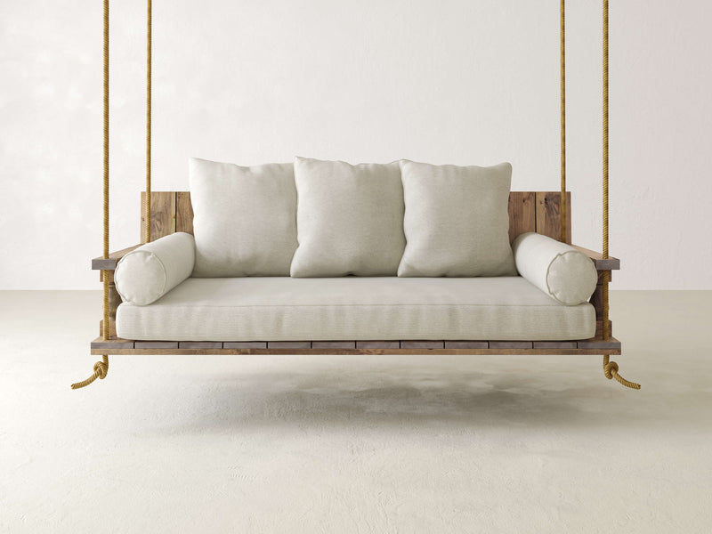 The Maisie Porch Swing by James+James is a handcrafted swing suspended from the ceiling by sturdy ropes, featuring a wooden frame and beige cushioning. This swing bed includes three large back pillows and two cylindrical armrest pillows, all in matching beige fabric. Set in a minimalist space with light-colored walls and floors, it ensures maximum comfort.