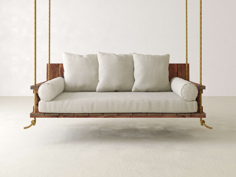 The James+James Maisie Porch Swing - Tuscany, featuring beige cushions, is suspended by ropes in a minimalist room with light-colored walls and floor. The swing includes three large square pillows and two round armrest cushions, creating a cozy and inviting seating area.