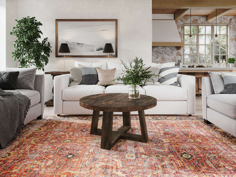 A cozy living room features two matching white sofas, a Rae Coffee Table in Tobacco by James+James at the center, and a colorful patterned rug. The back wall is adorned with framed artwork and two black wall lamps. A potted plant stands in the corner, with large windows letting in natural light.