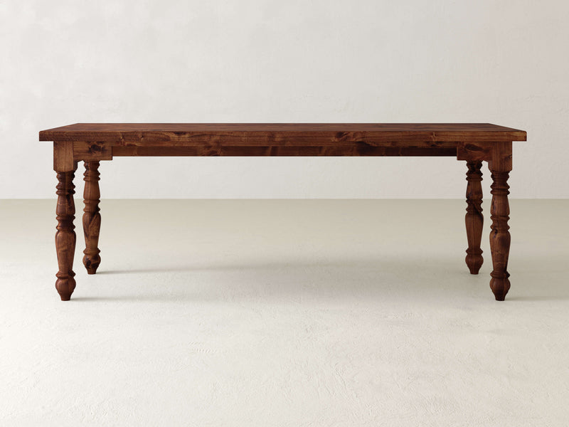 A hardwood dining table with intricately carved legs resembles a Turned Leg Desk by James+James, set elegantly on a light-colored floor against a plain white wall.