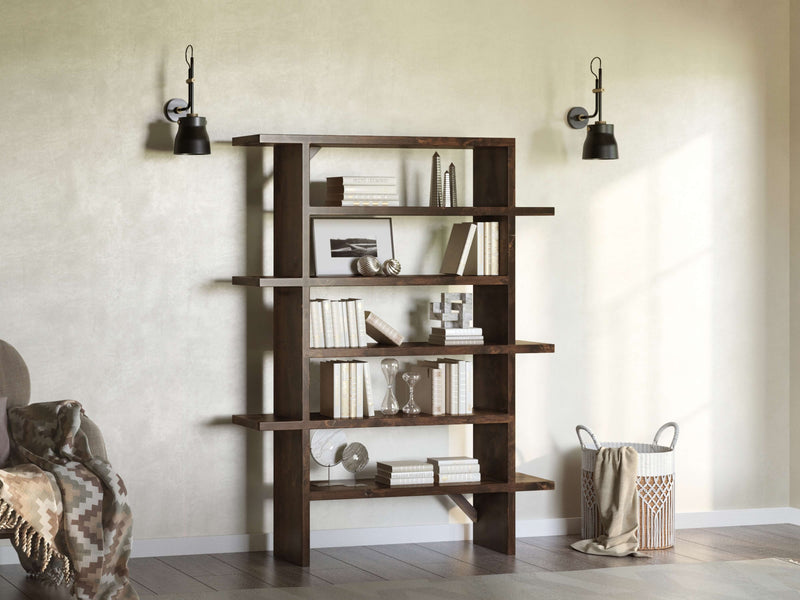 A James+James Unique Shelf, a modern handmade wooden bookshelf with books and decor, stands against a light wall flanked by two wall-mounted lamps. Nearby, a cozy chair draped with a patterned blanket sits next to a woven basket.