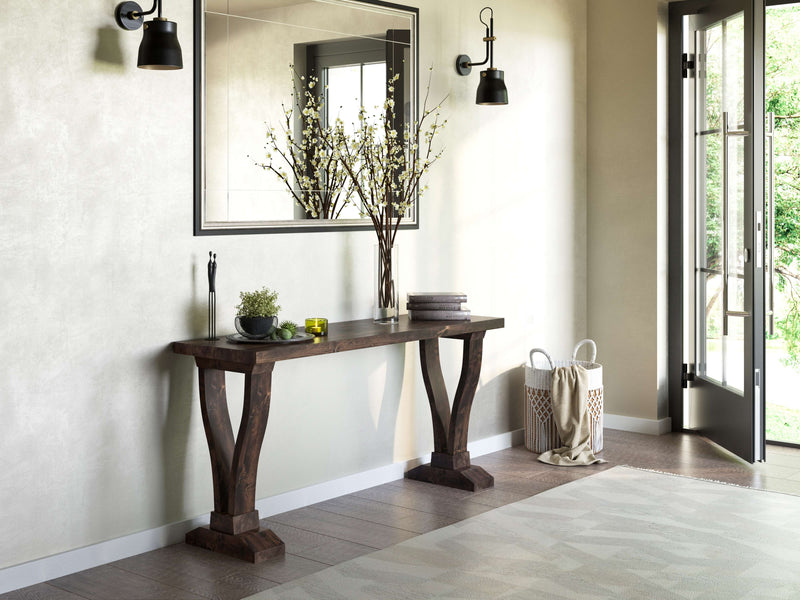 A modern, minimalist entryway features the Vera Sofa Table - Tobacco from James+James adorned with a vase of tall branches, a small green candle, and stacked books. Two black wall sconces flank a large mirror above the table. A light-filled doorway opens to a lush green garden.