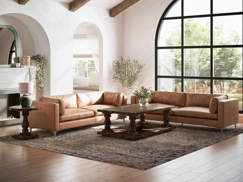 A spacious living room features two brown leather sofas arranged around a large wooden coffee table. A patterned rug covers the wooden floor, and a Vivien Side Table in Tobacco by James+James completes the setup. Arched windows and doorways allow natural light to flood the space, while a potted plant near the window adds a touch of greenery.