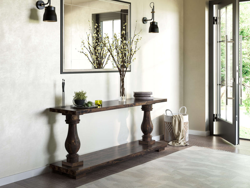 A minimalist hallway features the Vivien Sofa Table in Tobacco from James+James, showcasing its rustic wooden charm with ornate legs. The table holds a tall vase filled with branches, a small tray of decorative items, and a stack of books. A large mirror hangs above the table, while wall-mounted sconces illuminate the space. An open door leads to a lush green garden.