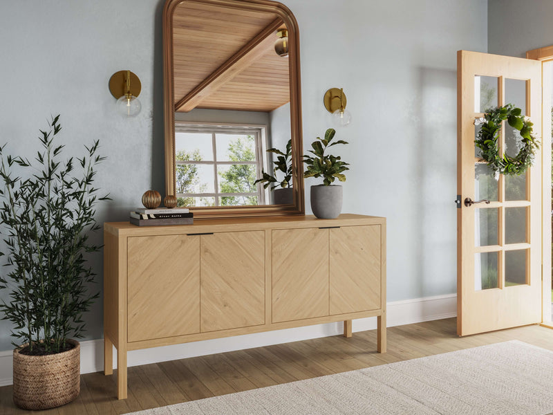 A modern entryway features a White Oak Buffet in a satin finish by James+James with four doors, a large arched mirror above, two plants (one potted on the buffet and one in a basket on the floor), and a wreath-adorned glass door. The space is illuminated by two wall sconces and natural light.