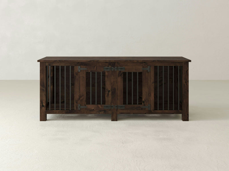 The Double Dog Kennel - Tobacco by James+James is a rustic wooden sideboard with a dark finish. It features two large central doors adorned with vertical metal bars and sturdy hinges. The piece stands on four legs and has a flat top surface, making it ideal for storage or display in a living or dining room.