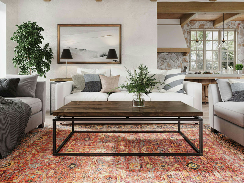 A modern living room features a white sofa, gray armchairs, and the Floating Top Coffee Table - Tobacco from James+James on an ornate red rug. Behind the sofa, a painting and two black table lamps decorate the wall. A green plant stands to the left, and the background reveals a spacious kitchen.