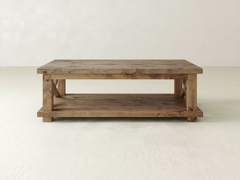 The Carter X Coffee Table by James+James is a rustic wooden piece with a rectangular shape and a lower shelf for additional storage. It features a harvest wheat finish and sturdy legs, reflecting a simple, minimalist design. The background includes a plain, light-colored wall and floor.